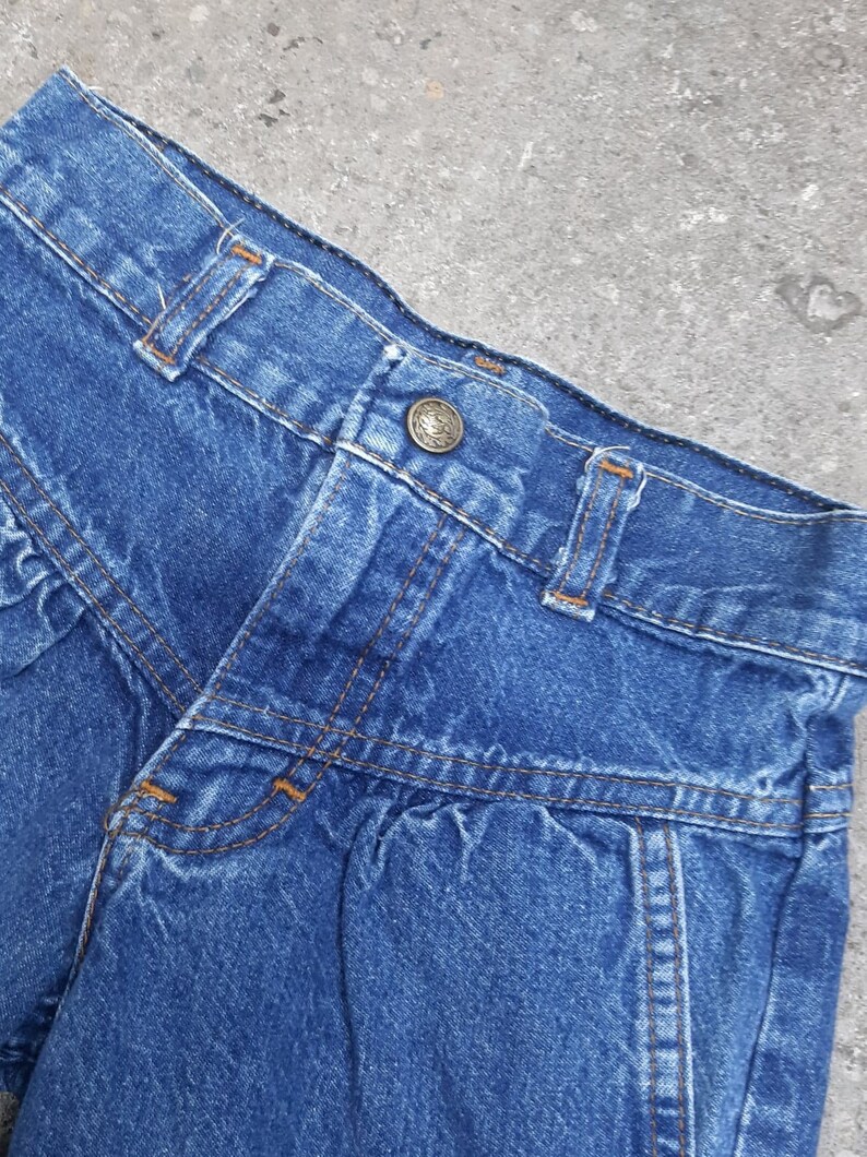 Puede incluir: Primer plano de unos pantalones cortos vaqueros azules. Los pantalones cortos tienen cierre de bot&oacute;n, trabillas y costuras decorativas. El denim tiene un aspecto ligeramente descolorido, lo que sugiere un estilo vintage. Los pantalones cortos son una prenda cl&aacute;sica e informal.