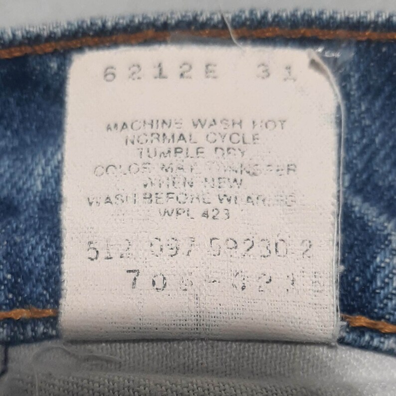 Puede incluir: Primer plano de una etiqueta blanca cosida a unos vaqueros de mezclilla azules. La etiqueta muestra instrucciones de lavado y c&oacute;digos de producto. Los vaqueros son de color azul claro.