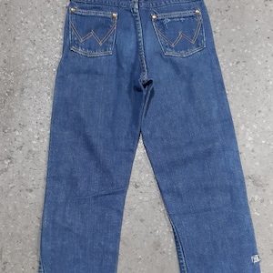May include: Blue denim jeans with the Wrangler logo on the back pockets. The jeans have a classic five-pocket design and a straight leg cut. The back pockets are stitched with a distinctive "W" pattern. The jeans are made of a durable denim fabric.