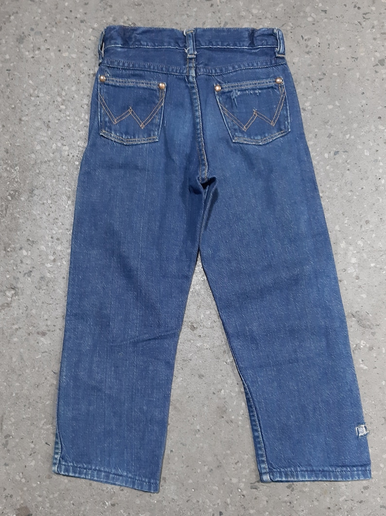 May include: Blue denim jeans with the Wrangler logo on the back pockets. The jeans have a classic five-pocket design and a straight leg cut. The back pockets are stitched with a distinctive "W" pattern. The jeans are made of a durable denim fabric.