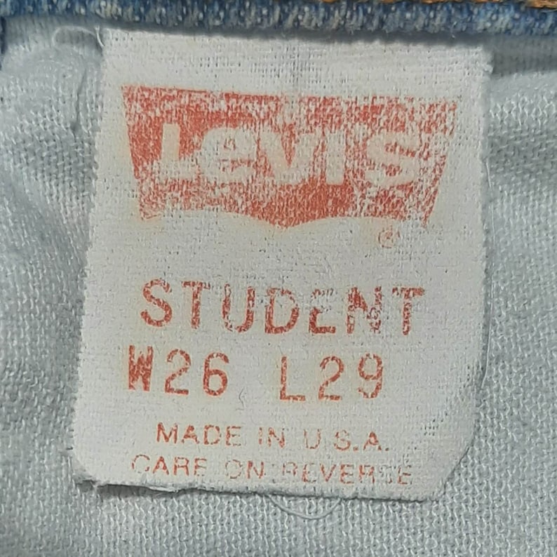 Puede incluir: Primer plano de una etiqueta blanca con letras rojas y un logotipo rojo. La etiqueta dice "LEVI'S STUDENT W26 L29 MADE IN U.S.A. CARE ON REVERSE". La etiqueta est&aacute; cosida en denim azul claro.