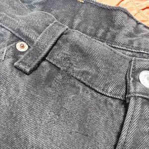 May include: Close-up of a pair of black denim jeans. The image highlights the details of the waistband, belt loop, and button closure. The denim has a textured appearance with visible stitching and a metal button.
