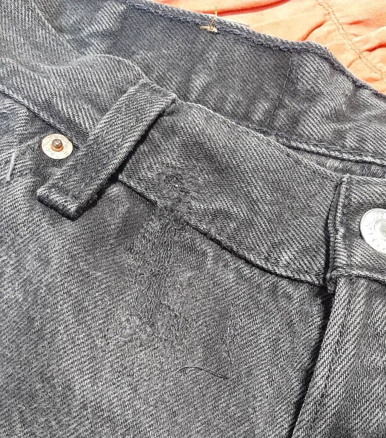 May include: Close-up of a pair of black denim jeans. The image highlights the details of the waistband, belt loop, and button closure. The denim has a textured appearance with visible stitching and a metal button.