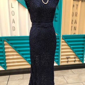 K&ouml;nnte beinhalten: Ein marineblaues Kleid im Vintage-Stil mit einem strukturierten, offen gewebten Design. Das Kleid hat einen V-Ausschnitt, Cap-&Auml;rmel und eine taillierte Taille mit passendem G&uuml;rtel. Eine Perlenkette schm&uuml;ckt die B&uuml;ste. Das Kleid wird auf einem schwarzen Mannequin-St&auml;nder pr&auml;sentiert.