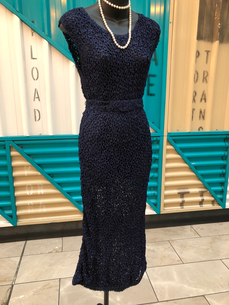 K&ouml;nnte beinhalten: Ein marineblaues Kleid im Vintage-Stil mit einem strukturierten, offen gewebten Design. Das Kleid hat einen V-Ausschnitt, Cap-&Auml;rmel und eine taillierte Taille mit passendem G&uuml;rtel. Eine Perlenkette schm&uuml;ckt die B&uuml;ste. Das Kleid wird auf einem schwarzen Mannequin-St&auml;nder pr&auml;sentiert.