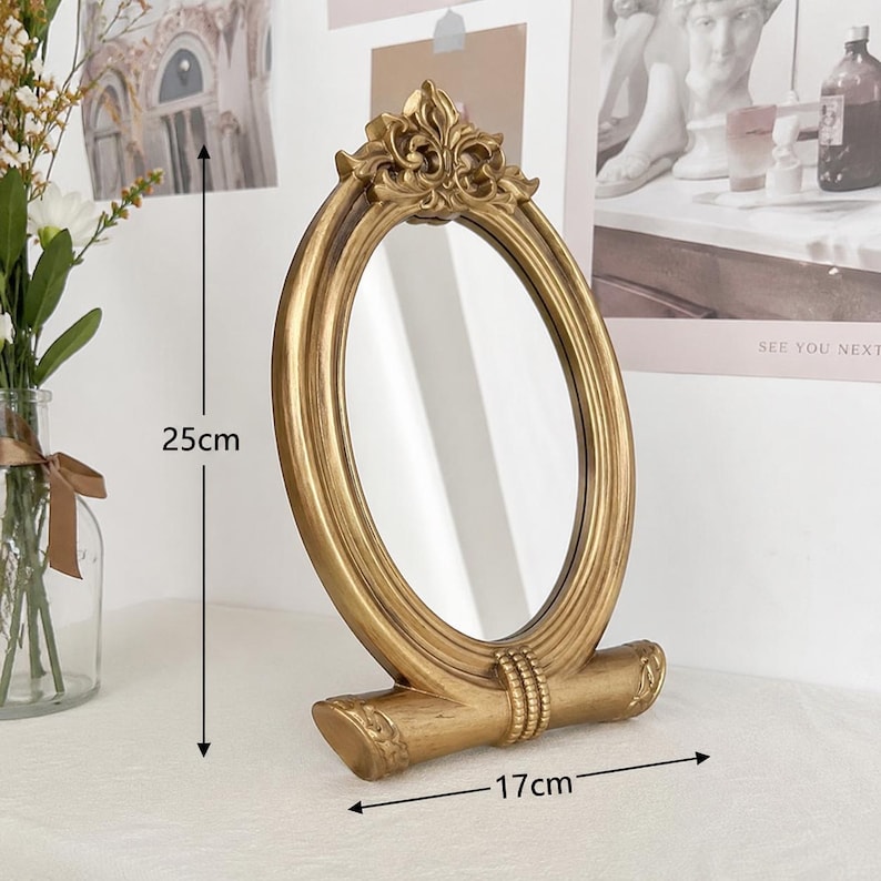 Może przedstawiać: Owalne lusterko stołowe ze złotą ramą ozdobioną ornamentami u g&oacute;ry i poziomą podstawą. Lusterko ma 25 cm wysokości i 17 cm szerokości, co jest zaznaczone czarnymi strzałkami oraz tekstem nadrukowanym bezpośrednio na obrazie.