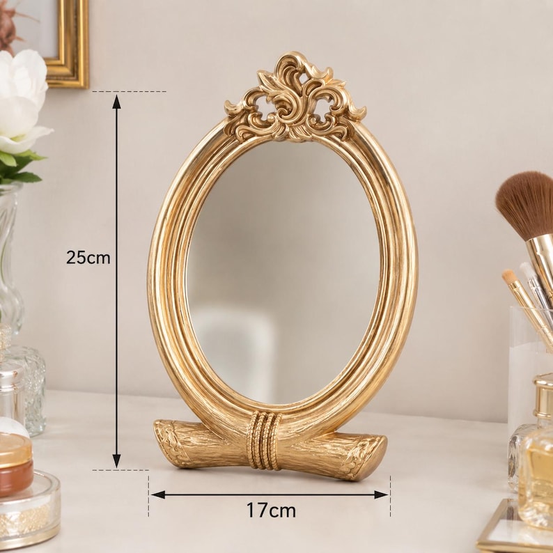Może przedstawiać: Owalne lusterko stołowe ze złotą ramą ozdobioną ornamentami u g&oacute;ry i teksturowaną podstawą. Lusterko ma 25 cm wysokości i 17 cm szerokości, co jest zaznaczone za pomocą widocznych na zdjęciu linii pomiarowych oraz odpowiednich etykiet.