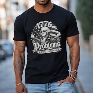May include: Black t-shirt with a graphic of George Washington in sunglasses, and the text "1776 Problems But England Ain't One." The design incorporates an American flag and laurel wreaths, conveying a patriotic message.