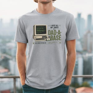 Puede incluir: Una camiseta gris jaspeado con un gráfico de un ordenador beige vintage y un disquete. El texto dice: I KEEP ALL MY JOKES IN A DAD-A-BASE, DAD-A-BASE SYSTEMS, INC. STORING DAD JOKES SINCE 1984. El diseño utiliza letras de color negro y verde.