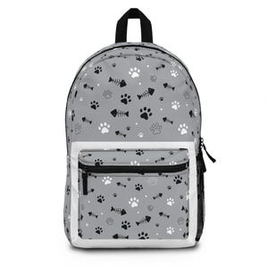May include: A gray backpack featuring a repeating pattern of black and white paw prints, fish skeletons, and small dots. The bag includes a front zippered pocket with a white border, a black top handle, and a black mesh side pocket for storage.