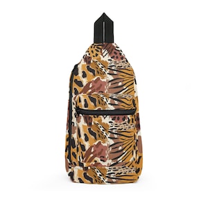 May include: A sling backpack featuring a multi-colored animal print pattern in shades of mustard yellow, brown, black, and cream. The bag has a single black top handle, a vertical side zipper, and a smaller horizontal zippered front pocket.