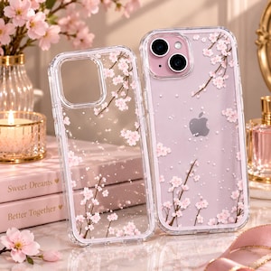 May include: Two clear phone cases featuring a delicate cherry blossom branch pattern with small pink flowers and white speckles. One case is empty, while the other is fitted onto a light purple smartphone with a dual camera lens and Apple logo.