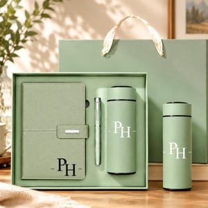 Op de afbeelding: Een saliegroene cadeauset bestaande uit een notitieboek met magnetische sluiting, een bijpassende balpen en twee cilindervormige geïsoleerde bekers. Het notitieboek en beide bekers zijn gepersonaliseerd met het monogram PH in schreeflettertype.