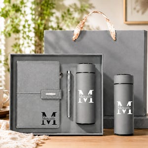 Op de afbeelding: Een grijze cadeauset met een notitieboek met magnetische sluiting, een zilveren pen en twee cilindrische vacuümgeïsoleerde flessen. Elk item is gepersonaliseerd met de letter M en de naam Metcalfe. De set wordt gepresenteerd in een grijze doos en tas.