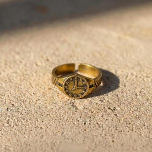 May include: A gold-colored ring with a watch face design. The ring features a round watch face with black accents and a band designed to resemble a watch strap. The hands of the watch are positioned at 2:00.