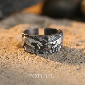 Può includere: Anello argentato con fascia strutturata raffigurante due mani che si tendono l'una verso l'altra. L'anello è appoggiato sulla sabbia, con la scritta "ronas." in basso. Un gioiello unico.