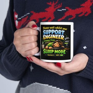May include: White ceramic mug with the text "Please wait whilst your SUPPORT ENGINEER resumes from SLEEP MODE". The mug features a graphic of a person sleeping at a computer. The mug is held by a person wearing a dark blue sweater.