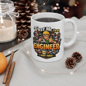 Puede incluir: Taza de cerámica blanca con el texto "Trust Me, I'm an ENGINEER" y un dibujo de un ingeniero. El ingeniero lleva un casco amarillo y hace una señal de aprobación. La taza contiene un líquido oscuro.