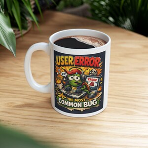 May include: White ceramic coffee mug with a black interior, featuring a colorful cartoon design. The design includes the text "USER ERROR" and "THE MOST COMMON BUG", along with a cartoon turtle character wearing headphones and using a laptop. The mug is on a wooden surface.