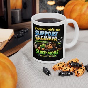 Könnte beinhalten: Weiße Keramiktasse mit schwarzer Grafik, die lautet: "Please wait whilst your SUPPORT ENGINEER resumes from SLEEP MODE." Die Tasse hat einen Henkel und ist mit einer dunklen Flüssigkeit gefüllt. Der Hintergrund zeigt Halloween-Dekorationen.