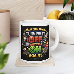 Może przedstawiać: Biały ceramiczny kubek z kolorową grafiką i napisem "Have you tried TURNING IT OFF AND ON AGAIN?". Wzór przedstawia postać z kreskówki z laptopem, zdenerwowaną obudowę komputera, filiżankę kawy oraz różne ikony związane z technologią.