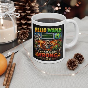 Op de afbeelding: Witte keramische mok met een zwarte afbeelding. De afbeelding toont de tekst "HELLO WORLD" in geel en groen, een screenshot met "print("Hello, World")" en cartoonmonsters. De zin "IS WHERE IT ALL WENT WRONG" is ook afgebeeld.