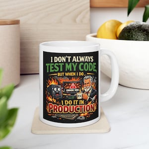 May include: White ceramic mug with a humorous design. The mug features the text "I DON'T ALWAYS TEST MY CODE, BUT WHEN I DO... I DO IT IN PRODUCTION" with graphics of a computer, error message, and a developer. The mug is on a beige coaster.