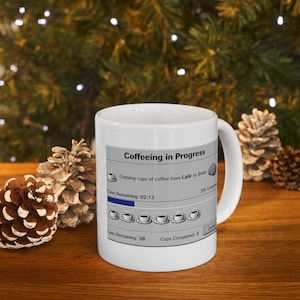 May include: White ceramic coffee mug with the text "Coffeeing in Progress" and a progress bar graphic. The mug features a humorous design of coffee cups copying from "Café to Brain". The mug is on a wooden surface.