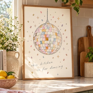 May include: A framed wall art print featuring a central disco ball illustration with pastel-colored square tiles, surrounded by small colorful confetti shapes and starbursts, with the text "this kitchen is for dancing" written below.