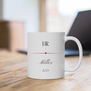 Könnte beinhalten: Ein weißer Keramikbecher mit dem schwarzen Text MR. oben, gefolgt von einer dünnen horizontalen Linie mit einem kleinen roten Herz in der Mitte. Unter der Linie ist der Name Miller in einer Schreibschrift gedruckt, mit der Jahreszahl 2025 unten.