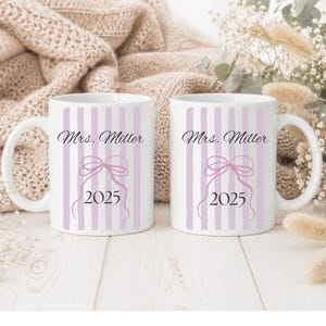 Könnte beinhalten: Zwei weiße Keramiktassen mit einem vertikalen hellvioletten Streifenmuster. Jede Tasse zeigt den Text Mrs. Miller in schwarzer Schreibschrift, eine hellviolette Strichzeichnung einer Schleife und das Jahr 2025 in schwarzer Serifenschrift.