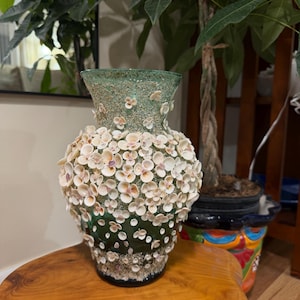 May include: A decorative vase with a dark green base and a light green top. The vase is adorned with small, white seashells, creating a textured, organic appearance. The vase sits on a wooden surface.
