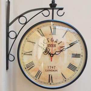 May include: A double-sided, vintage-style wall clock with a black metal frame and bracket. The clock face is cream-colored with black Roman numerals and the words "VICTORIA STATION 1747 LONDON". The clock hands are black with a red second hand.