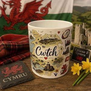 Könnte beinhalten: Eine weiße Keramiktasse mit einer Karte von Wales, dem Wort "Cwtch" und Illustrationen von Sehenswürdigkeiten. Die walisische Flagge ist im Hintergrund. Ein Schieferuntersetzer mit "CYMRU" ist ebenfalls sichtbar.