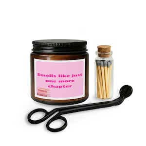 Puede incluir: Un tarro de vela marrón con tapa negra y una etiqueta rosa que dice "Smells like just one more chapter". También hay una pequeña botella de vidrio de cerillas y un recortador de mecha negro.