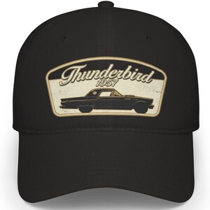 Puede incluir: Una gorra de béisbol negra con un parche gráfico centrado. El parche tiene un fondo color crema con la silueta negra de un coche Thunderbird de 1957 y el texto Thunderbird 1957 en una fuente cursiva estilizada, todo en un borde oscuro.