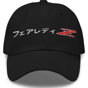 May include: Black baseball cap featuring white Japanese text and a red stylized "Z" logo. The cap has a curved peak and an adjustable fabric strap. The hat is made of a soft, durable material.