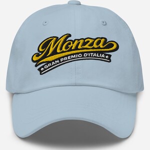 May include: Light blue baseball cap featuring the word "Monza" in a yellow and black script, with "Gran Premio D'Italia" below. The cap has a curved brim and a classic six-panel design, suitable for casual wear.