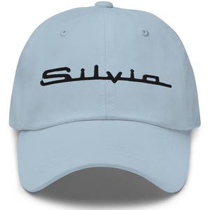 May include: Pale blue baseball cap featuring the word "Silvia" embroidered in black. The cap has a curved peak and an adjustable strap at the rear.