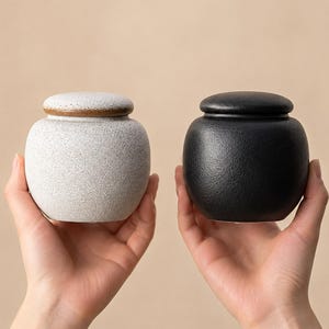 May include: Two small ceramic jars with lids, one featuring a speckled off-white finish and the other a solid matte black finish. Both containers have a rounded, bulbous shape with a smooth texture and a distinct rim where the lid meets the base.