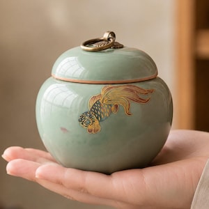 May include: A small, round, light green ceramic jar with a matching lid featuring a metallic ring handle. The jar is decorated with a detailed, colorful illustration of a goldfish with orange, yellow, and black scales swimming on its side.