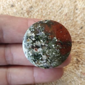 May include: A circular polished gemstone cabochon featuring a mottled pattern of deep forest green, earthy olive, and vibrant brick red inclusions with small white crystalline flecks distributed across the smooth, rounded surface.