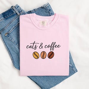 May include: A light pink t-shirt featuring the black text "cats & coffee" above three illustrated coffee beans in shades of yellow, light brown, and dark brown, paired with a pair of blue denim jeans.