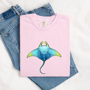 Könnte beinhalten: Ein hellrosa T-Shirt mit einer grafischen Illustration eines Mantarochens. Der Mantarochen ist in Blau-, Türkis- und Gelbtönen mit einem langen Schwanz dargestellt. Das T-Shirt hat einen Rundhalsausschnitt und ein sichtbares Markenetikett am Kragen.