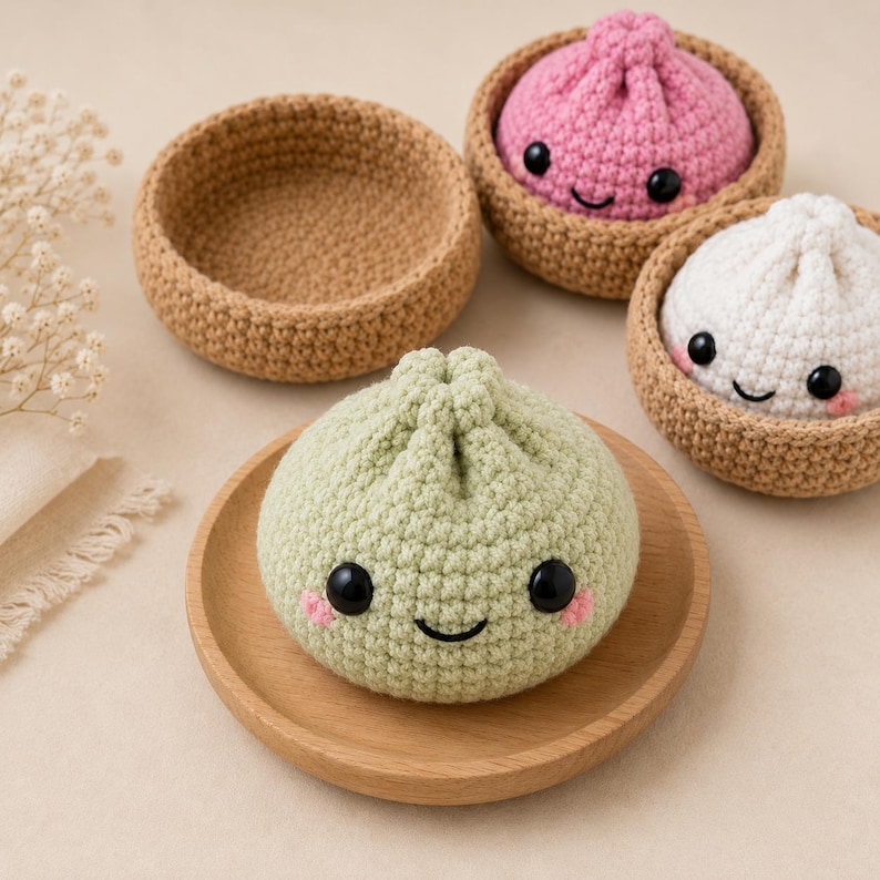 May include: Three crocheted dumpling toys with smiling faces and black eyes. One is light green on a wooden plate, one is pink in a brown crocheted bowl, and one is white in a brown crocheted bowl. An empty brown crocheted bowl sits nearby.