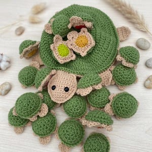 以下が含まれることがあります： 緑とベージュの色合いの手作りのカギ針編みのカメ。カメは、花のようなアクセントが付いた大きな渦巻きの甲羅と、周りにいくつかの小さなカメがあります。カメは糸でできており、柔らかく、質感のある外観をしています。
