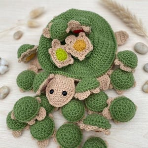 May include: A crochet turtle toy featuring a large green shell with a beige underside and head, adorned with several smaller green and beige baby turtles on top. The baby turtles have small yellow and light green accents on their shells.