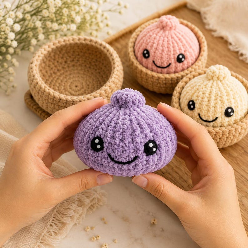 May include: A purple crocheted dumpling toy with a smiling face and black eyes, held in front of two other crocheted dumplings in tan baskets, one pink and one cream, all featuring rounded shapes and small top knots.