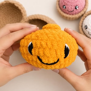 May include: A bright orange crocheted plush toy with a smiling face, black embroidered eyes, and a small top protrusion. The toy is made of soft yarn and is part of a set that includes other crocheted items in tan, pink, and white colors.