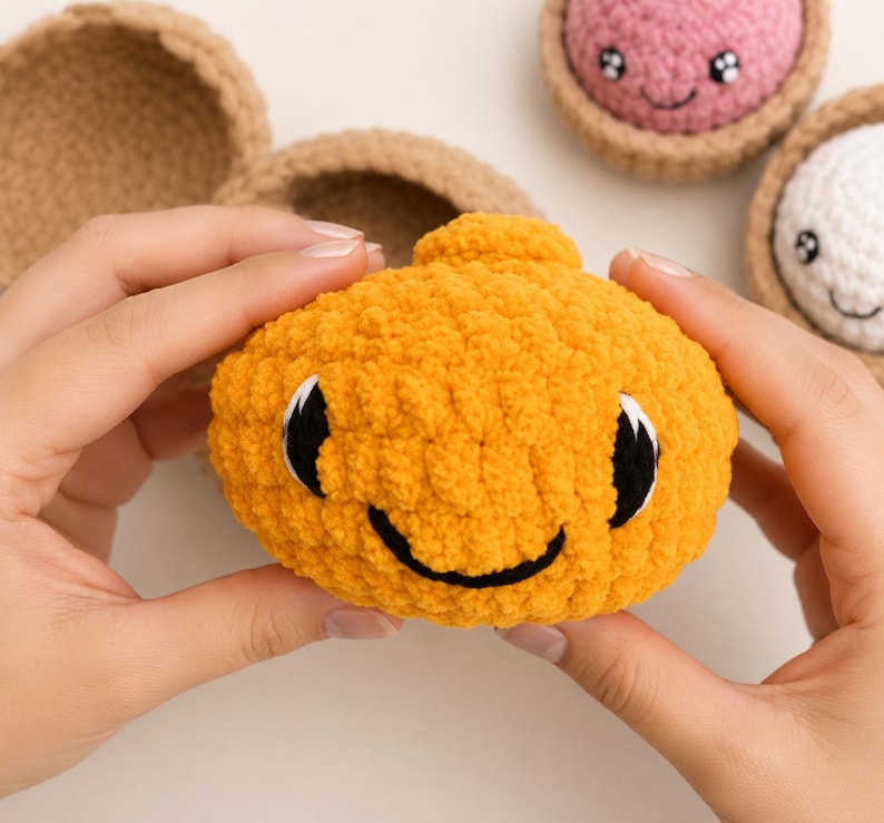 May include: A bright orange crocheted plush toy with a smiling face, black embroidered eyes, and a small top protrusion. The toy is made of soft yarn and is part of a set that includes other crocheted items in tan, pink, and white colors.